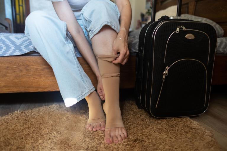Woman sitting on bed next to a suitcase, pulling up compression sock.